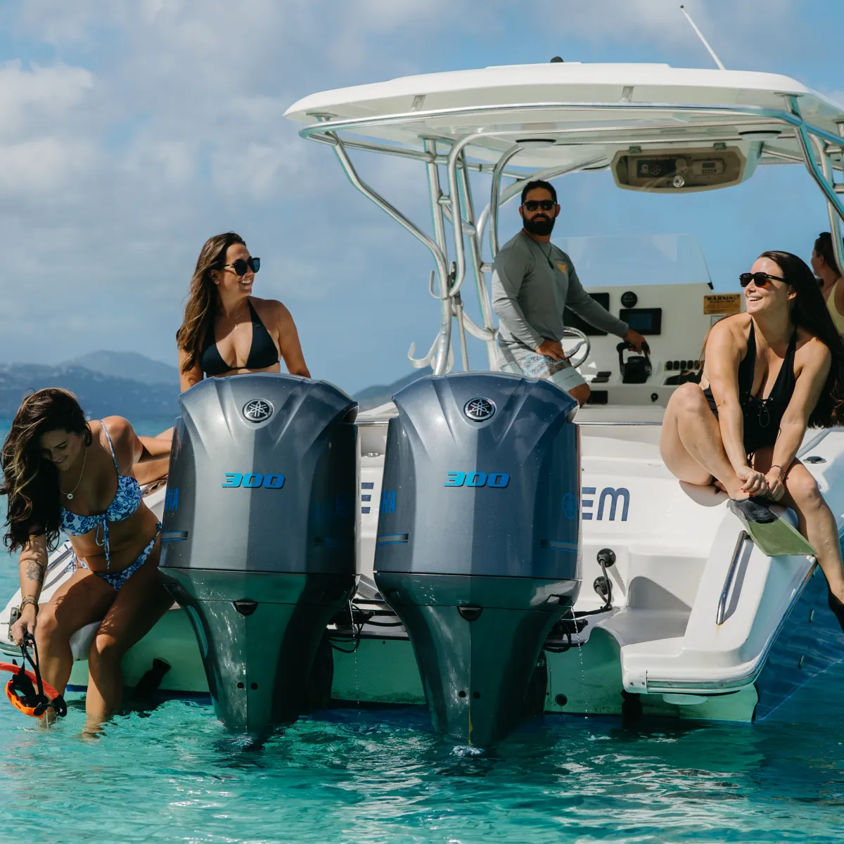 People on a boat in clear blue water, with two outboard motors and distant islands visible.