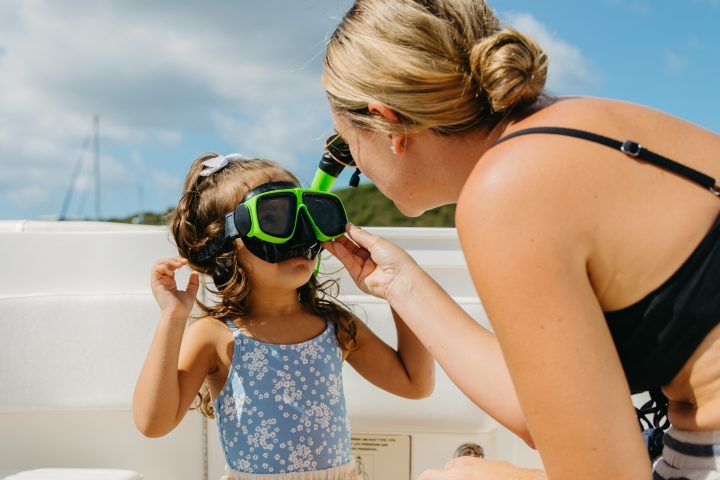 Woman helps young girl put on green snorkeling mask on a boat.