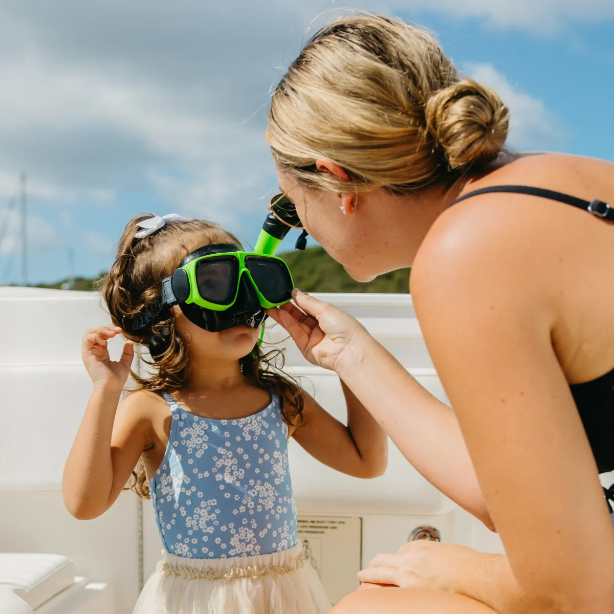 Woman helps young girl put on green snorkeling mask on a boat.